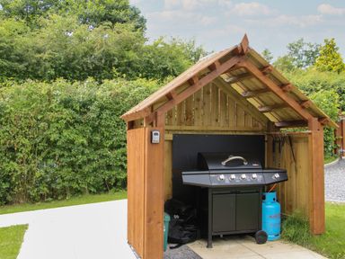 A gas barbecue grill under a wooden shelter with a propane tank outside in a garden at Jacobs Meadow Pod 2 in Minsterley near Chirbury