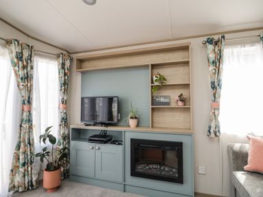 A living room with a TV on a cabinet and a fireplace with potted plants and curtains at Burlees Nest in Brigham near Great Broughton Cockermouth