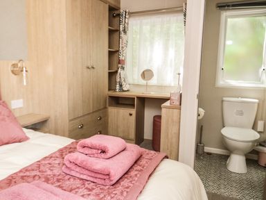 A bedroom with a bed with pink towels a wooden wardrobe a dressing table and a view of a bathroom with a toilet and a window at Burlees Nest in Brigham near Great Broughton Cockermouth
