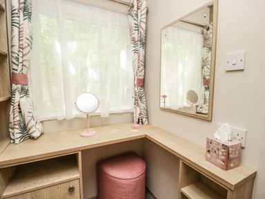 A corner dressing table with a round mirror, a small pink stool, floral curtains on a window, and a tissue box at Burlees Nest in Brigham near Great Broughton Cockermouth