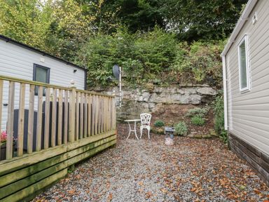 A small gravel patio area with a wooden railing and an outdoor table and chair set between two mobile homes at Burlees Nest in Brigham near Great Broughton Cockermouth