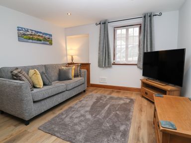 A living room with a gray sofa and cushions a wooden TV stand with a television a wooden corner cabinet a window with gray curtains and a floor lamp at Fryston Cottage in Keswick