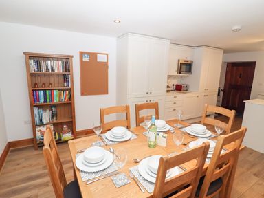 A dining area with a wooden table set for six and a bookshelf and kitchen cabinets in the background at Fryston Cottage in Keswick