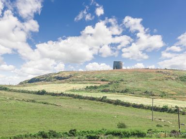 A stone tower on a grassy hill under a blue sky with scattered clouds at Cae Berllan in Mynytho near Pwllheli