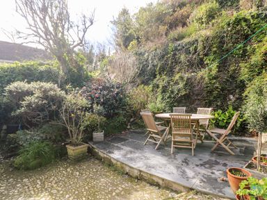 A garden patio with a round wooden table and six wooden chairs surrounded by plants and greenery at Widgeons Keel in Flushing