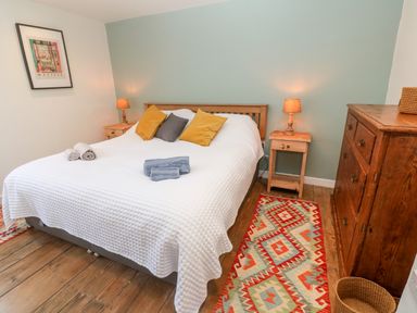 A bedroom with a double bed white bedspread and colorful pillows two bedside tables with lamps a wooden chest of drawers and a patterned rug on wooden floor at Widgeons Keel in Flushing