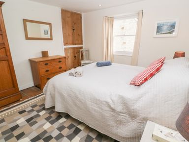 A bedroom with a bed covered in a white quilt and red checked pillow a wooden dresser mirror wooden wardrobe patterned rug and window at Widgeons Keel in Flushing