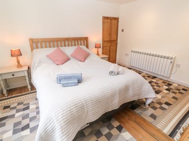 A bedroom with a bed with white bedding red checkered pillows bedside tables with lamps a wooden door and patterned rugs on a wooden floor at Widgeons Keel in Flushing