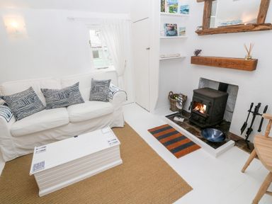 A living room with a white sofa with patterned cushions a white coffee table a wood stove fireplace with wood and tools and a wooden chair at The Little Net Loft in Mousehole near Penzance