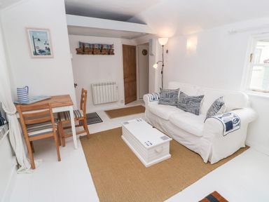 A living room with a white sofa with patterned cushions a wooden table with two chairs a white chest used as a coffee table and a floor lamp at The Little Net Loft in Mousehole near Penzance