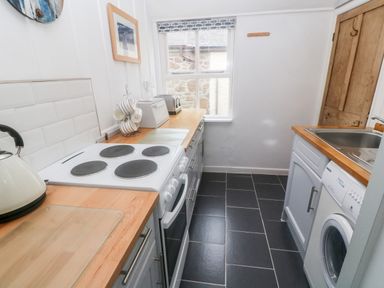 A narrow kitchen with an electric stove kettle toaster bread bin washing machine and sink at The Little Net Loft in Mousehole near Penzance