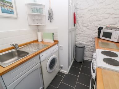 A small kitchen with a stainless steel sink washing machine stove microwave and trash bin at The Little Net Loft in Mousehole near Penzance