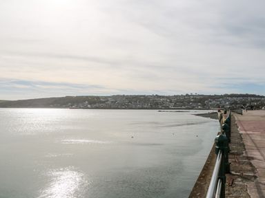 A waterfront promenade with railings along calm water and a town on the hillside in the distance at The Little Net Loft in Mousehole near Penzance