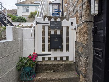 A white wooden gate with the number 5 and a mailbox at the bottom of stone steps with a potted plant next to it at The Little Net Loft in Mousehole near Penzance