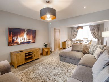 A living room with a large grey sofa a wooden TV stand a wall mounted TV showing a fireplace and a dining area with a wooden table and chairs at Family House in Weymouth