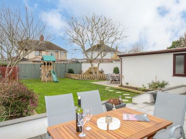 A backyard with a wooden table and chairs a children's playset with swings and slide and two houses in the background at Family House in Weymouth