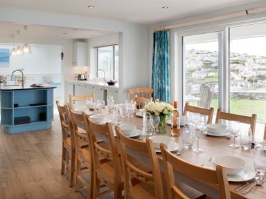 A dining area with a wooden table set with white dishes and glasses next to a kitchen with blue island and large windows at The White House in Polzeath