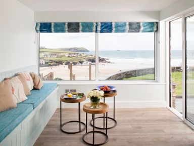 A seating area with a blue cushioned bench and three wooden round tables near large windows overlooking a beach at The White House in Polzeath