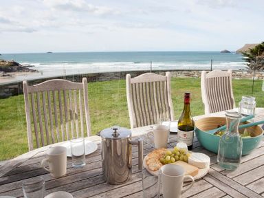 A wooden outdoor dining table with cups glasses a wine bottle cheese crackers grapes salad bowl and a carafe of water with chairs and an ocean view at The White House in Polzeath