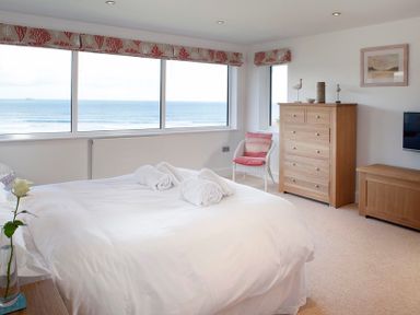 A bedroom with a large bed near windows overlooking the ocean with a wooden dresser and a chair at The White House in Polzeath