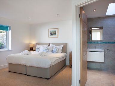 A bedroom with a double bed and pillows next to a window and an open door showing a bathroom with a sink and mirror at The White House in Polzeath