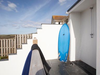 An outdoor shower area with a blue surfboard leaning against a white wall and wetsuits hanging on a wooden railing at The White House in Polzeath