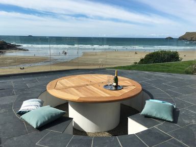 An outdoor patio with a round wooden table surrounded by built-in seating and cushions overlooking the beach and ocean at The White House in Polzeath