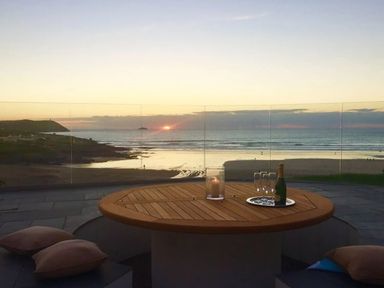 An outdoor seating area with a round wooden table cushions a candle bottle and glasses with an ocean view at sunset at The White House in Polzeath