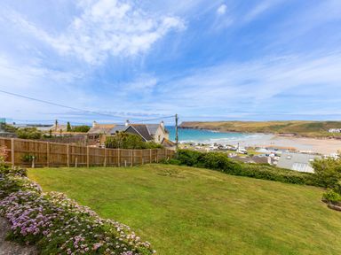 A grassy backyard with flowers along a stone path a wooden fence houses and a beach with sea and hills in the background at Zapadiah in Polzeath