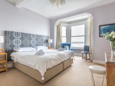 A bedroom with a large bed and headboard by a window with curtains and a blue armchair at 4 The Terrace in Rock