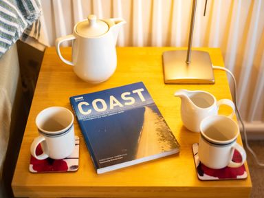 A wooden side table with a teapot two mugs a milk jug a lamp and a book titled Coast at Pencreek Rock