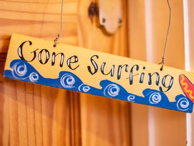 A wooden sign hanging on a wooden door that reads Gone Surfing with painted blue waves at Pencreek Rock
