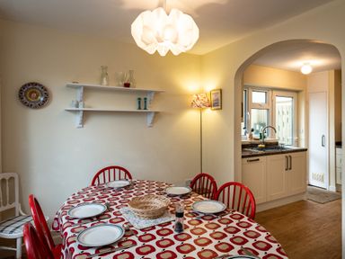 A dining area with a table covered by a tablecloth with a fruit pattern set with plates and cutlery red chairs surrounding the table and a kitchen visible through an archway at Pencreek in Rock