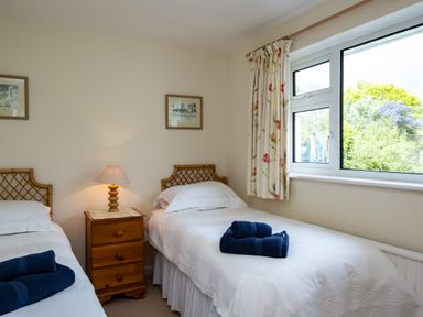 A bedroom with two single beds with white bedding and blue towels on each bed a wooden bedside table with a lamp and floral curtains on a window at Pencreek in Rock