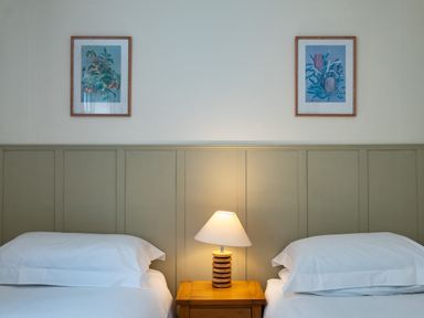 Two beds with white linens separated by a wooden nightstand with a lamp and two framed botanical prints on the wall above at Pencreek in Rock