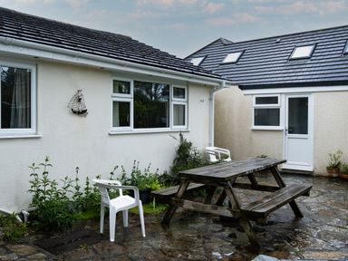 An outdoor patio area with a wooden picnic table and white plastic chairs near a single story building with white walls and windows at Pencreek in Rock