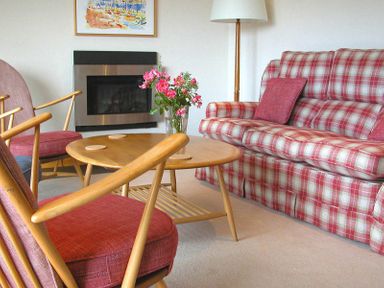 A living room with wooden armchairs a round wooden coffee table with flowers a plaid sofa and a wall-mounted fireplace at Rosevallen in Daymer Bay