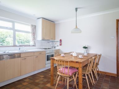 A kitchen with wooden cabinets a dining table with chairs and a window with floral curtains at Rosevallen in Daymer Bay
