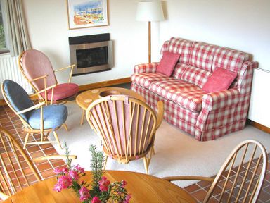 A living room with a red and white checkered sofa, wooden chairs with colored cushions, a round wooden table with flowers, a floor lamp, and a wall-mounted fireplace at Rosevallen in Daymer Bay