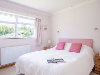A bedroom with a bed with pink pillows and a white cover near a window with curtains and wooden bedside tables at Rosevallen in Daymer Bay