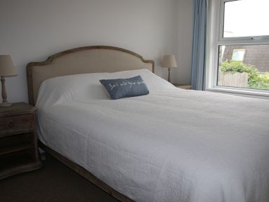 A bedroom with a bed covered in white bedding a blue pillow two bedside tables with lamps and a window with blue curtains at Slipway 10 in Rock
