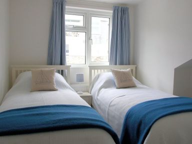 A bedroom with two single beds each with a sailboat pillow and blue throw with a window and blue curtains behind at Slipway 10 in Rock