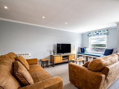 A living room with brown sofas a wooden TV stand a flat screen TV and a window seat with cushions at Slipway 15 in Rock