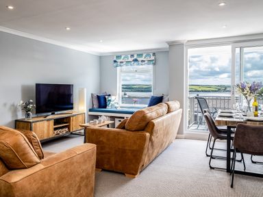 A living room with brown sofas a TV on a wooden stand a window seat with cushions a dining table set with chairs and flowers overlooking a balcony with a view at Slipway 15 in Rock
