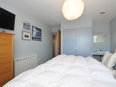 A bedroom with a bed covered in white bedding a wooden dresser television on the wall blue wardrobe doors a small sink with a mirror and paintings on the wall at Slipway 16 in Rock