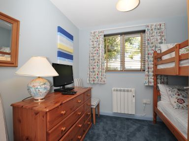 A bedroom with wooden bunk beds a wooden dresser with a lamp and a television a window with floral curtains and a chair at Slipway 16 in Rock