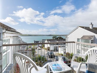A balcony with two white plastic chairs and a small table with books and a glass bottle overlooking a street and water at Slipway 19 in Rock