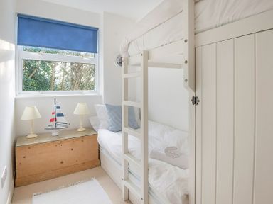 A bedroom with a white bunk bed two lamps and a model sailboat on a wooden chest under a window at Slipway 19 in Rock