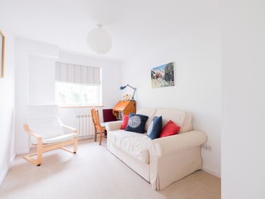 A small room with a beige sofa with cushions a wooden chair a white armchair a window with a striped blind and a wall painting at Slipway 21 in Rock