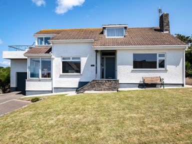 A white house with a tiled roof a front door a bench and a grassy lawn under a blue sky at Little Belz in Polzeath